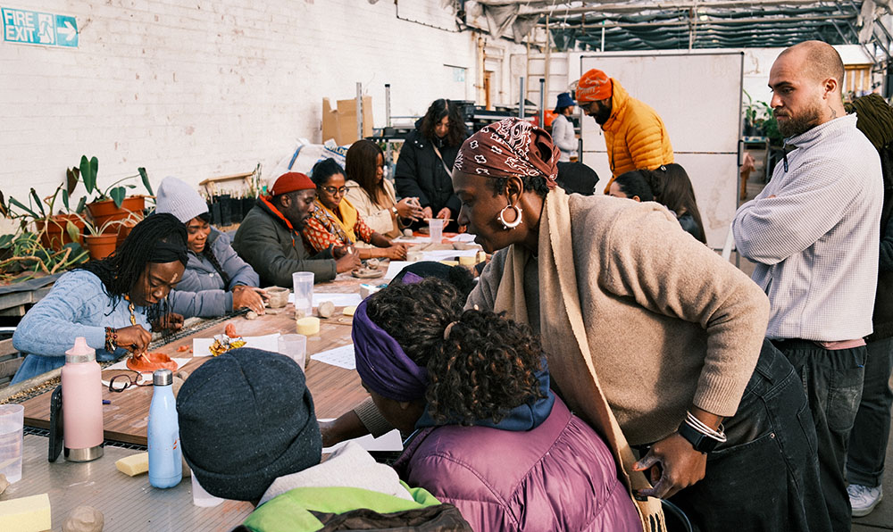 The image shows a group of people attending a workshop. The group are sat around a table and are taking part in a crafts workshop. We see a bright room in the background with plants. It looks like the day might be cold as lots of the group are wearing coats and hats.