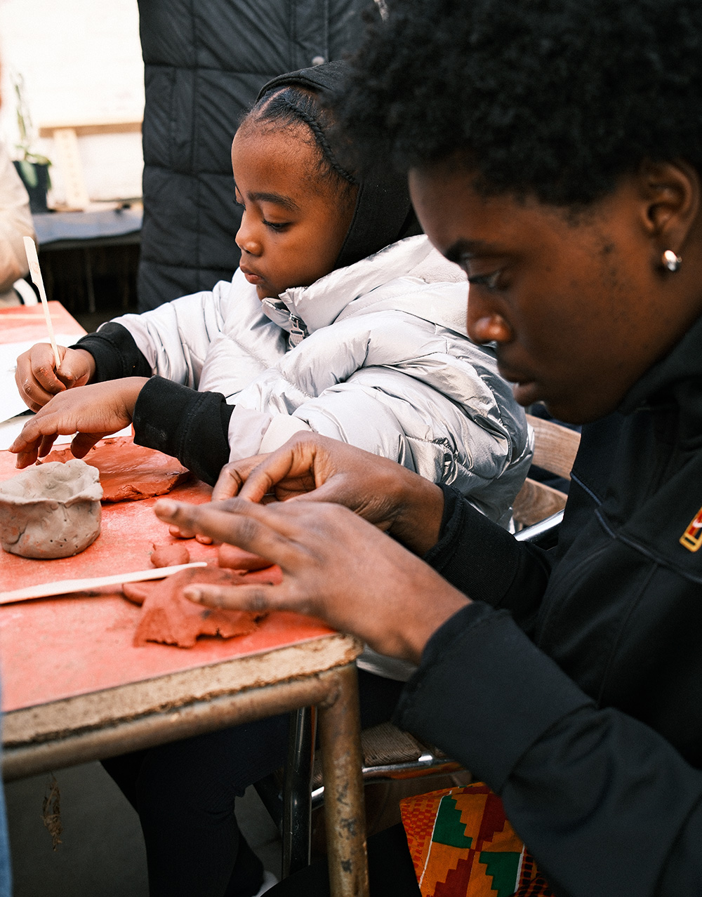The image shows a young child and an older young person taking part in a crafts workshop. They are sat next to each other and are concentrating on the task in front of them. It looks like they are carving or drawing into clay.