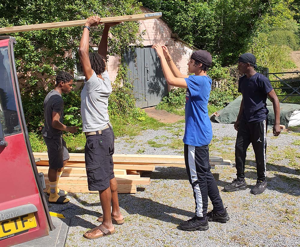 The image shows four young people working together to pack planks of wood into a red car. It is a sunny day and in the background we can see a building, lots of trees and greenery and a gate.