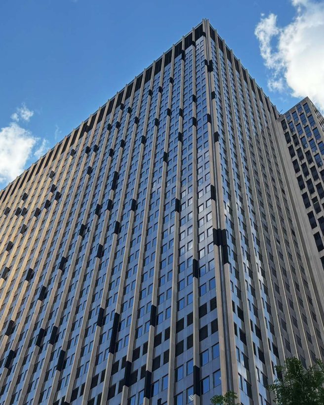 The image shows an upwards view of a large skyscraper. There is bright blue sky and white clouds.