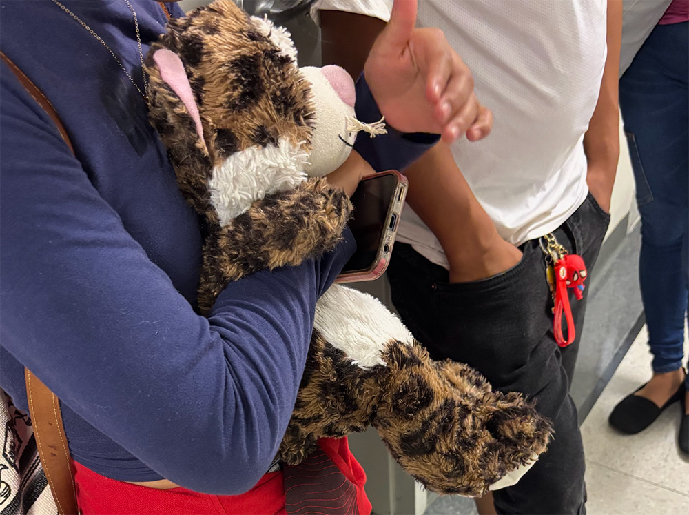 The image shows a young person in a long sleeve blue top holding onto a large teddy and a mobile phone. Next to them is a figure stood with a white shirt and black jeans. In the image you can't see the figures faces.