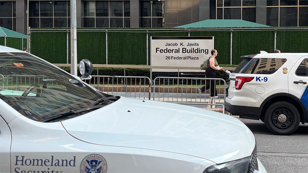 In the image we see the street with the sign Federal Building. There is a white and blue car on the right hand side of the image and to the left is a grey vehicle with the words, Homeland Security written on its side.