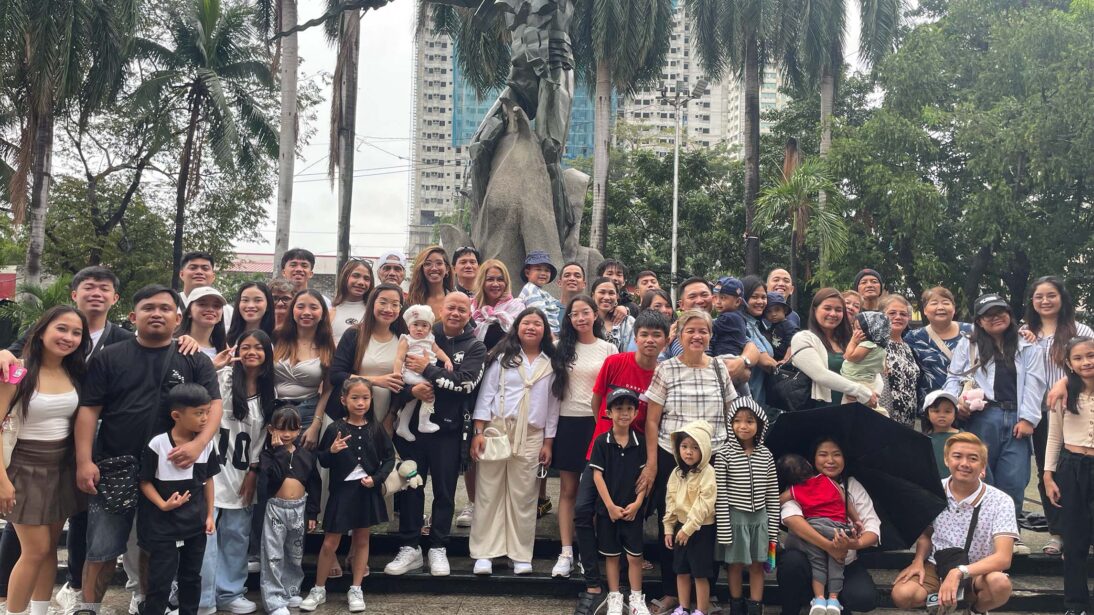The image shows a picture of Mitzi surrounded by her family in front of a statue in the plaza. They are all crowded around facing the camera to take a family photo.