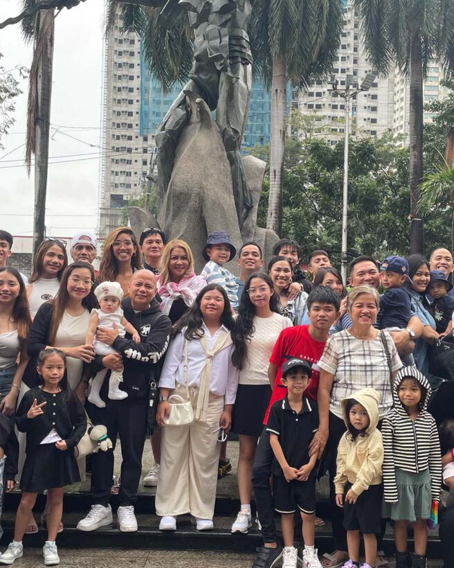 The image shows a picture of Mitzi surrounded by her family in front of a statue in the plaza. They are all crowded around facing the camera to take a family photo.