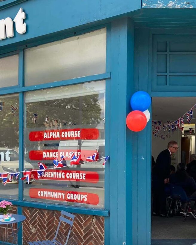 The image shows The Point in Lewisham. We see the entrance, with the door open. The building is painted blue and there are big windows with red signs on them. The sign, The Point is white and at the top of the building.