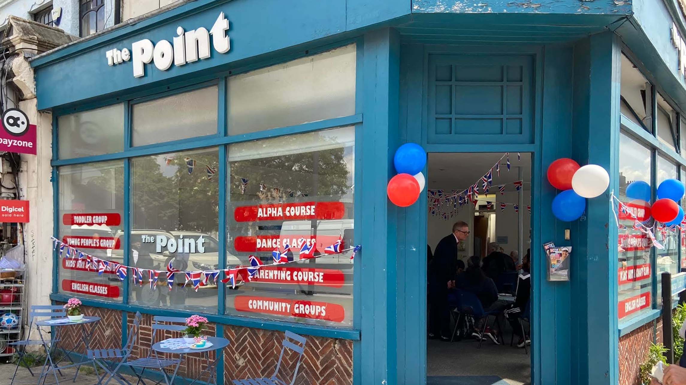The image shows The Point in Lewisham. We see the entrance, with the door open. The building is painted blue and there are big windows with red signs on them. The sign, The Point is white and at the top of the building.