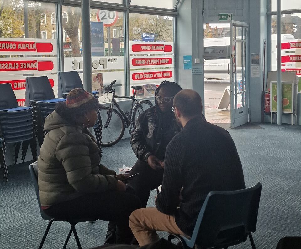 The image shows three people sat in a circle on plastic chairs in the centre of The Point. They are sat facing each other in discussion.