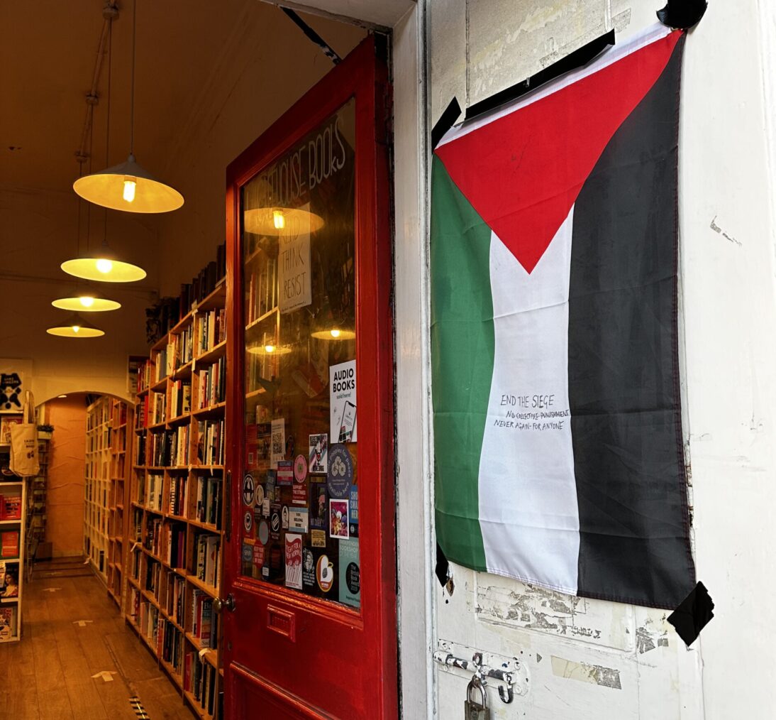 The image shows the entrance to the bookshop. In the foreground is a white wall next to the doorway and there is a Palestine flag hanging down. The door to the bookshop is red and inside we can see rows of bookshelves.