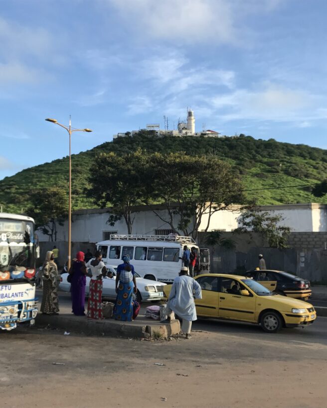 The image shows a green hill with a lighthouse on top of it. In the foreground we see a busy road with a bus and yellow taxis, alongside a group of five people waiting in the middle.