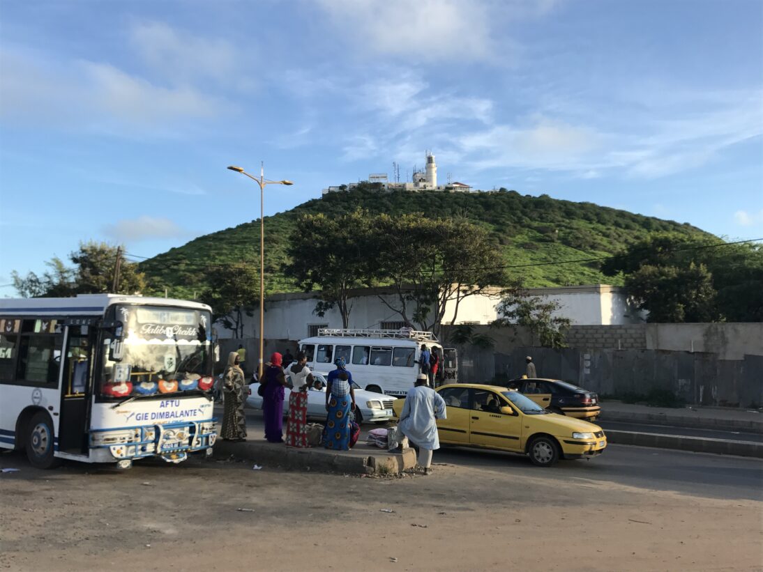 The image shows a green hill with a lighthouse on top of it. In the foreground we see a busy road with a bus and yellow taxis, alongside a group of five people waiting in the middle.