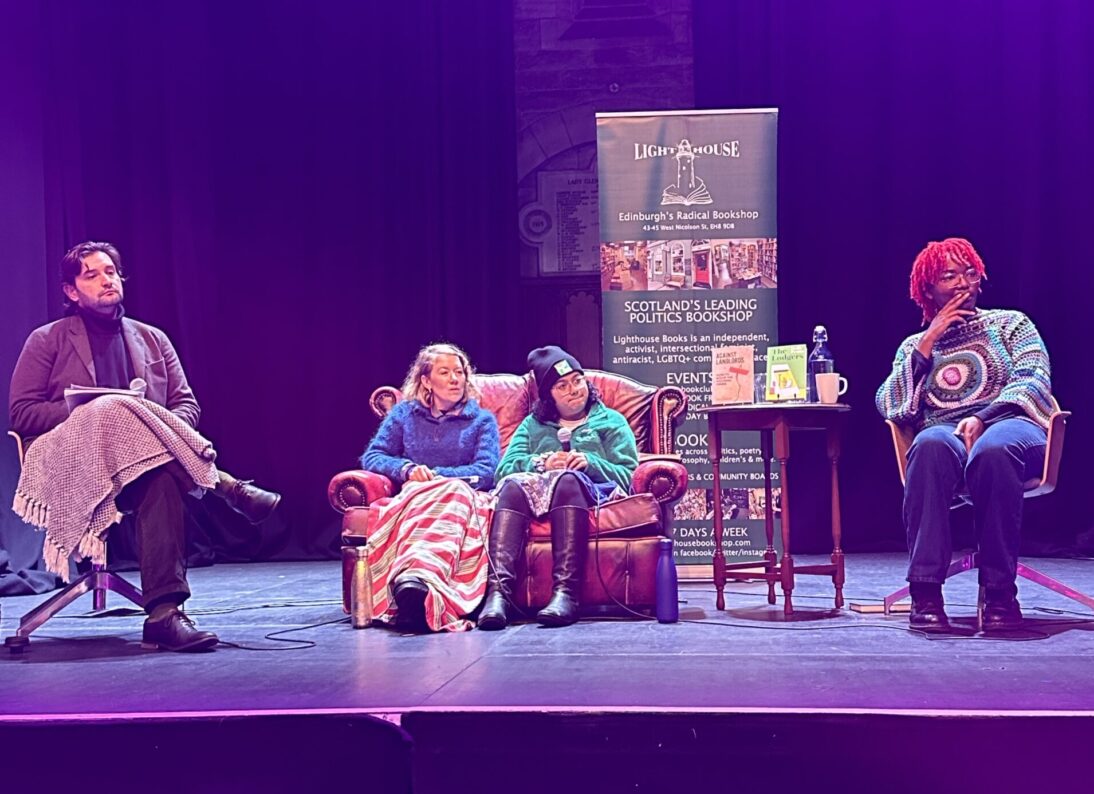 The image shows four people sat on a stage. In the centre are two people sat on a brown leather sofa looking outwards to the audience. To the left is a person sat on a taller chair looking out to the audience. They wear a black roll neck and a blazer. On the right hand side is the final person, also sat on a high chair and looking out to the audience. They are wearing a blue and purple patterned jumper.