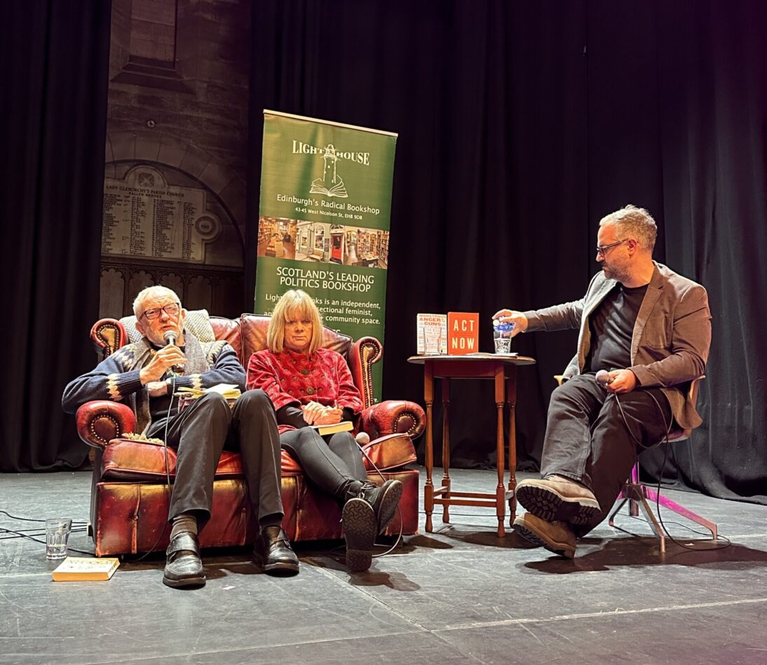 The image shows Jeremy Corbyn and Kate Pickett sat on a brown leather sofa on a stage. Jeremy is holding a microphone. There is one other person sat to the right, wearing a brown blazer.