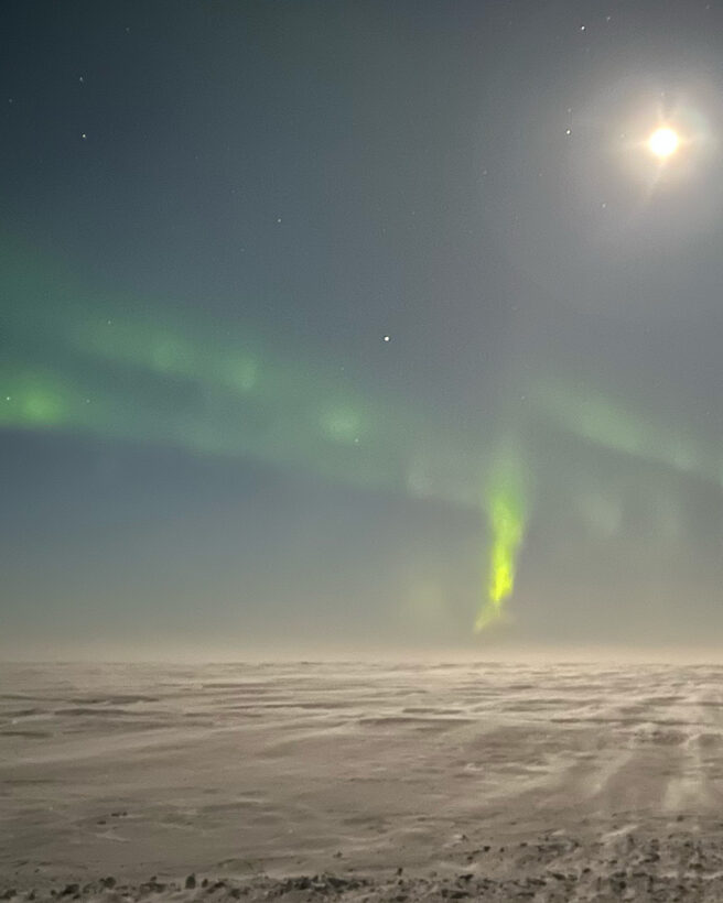 The image shows a landscape with a dark sky and a bright moon. There are wisps of the northern lights in green. The ground is covered in snow.