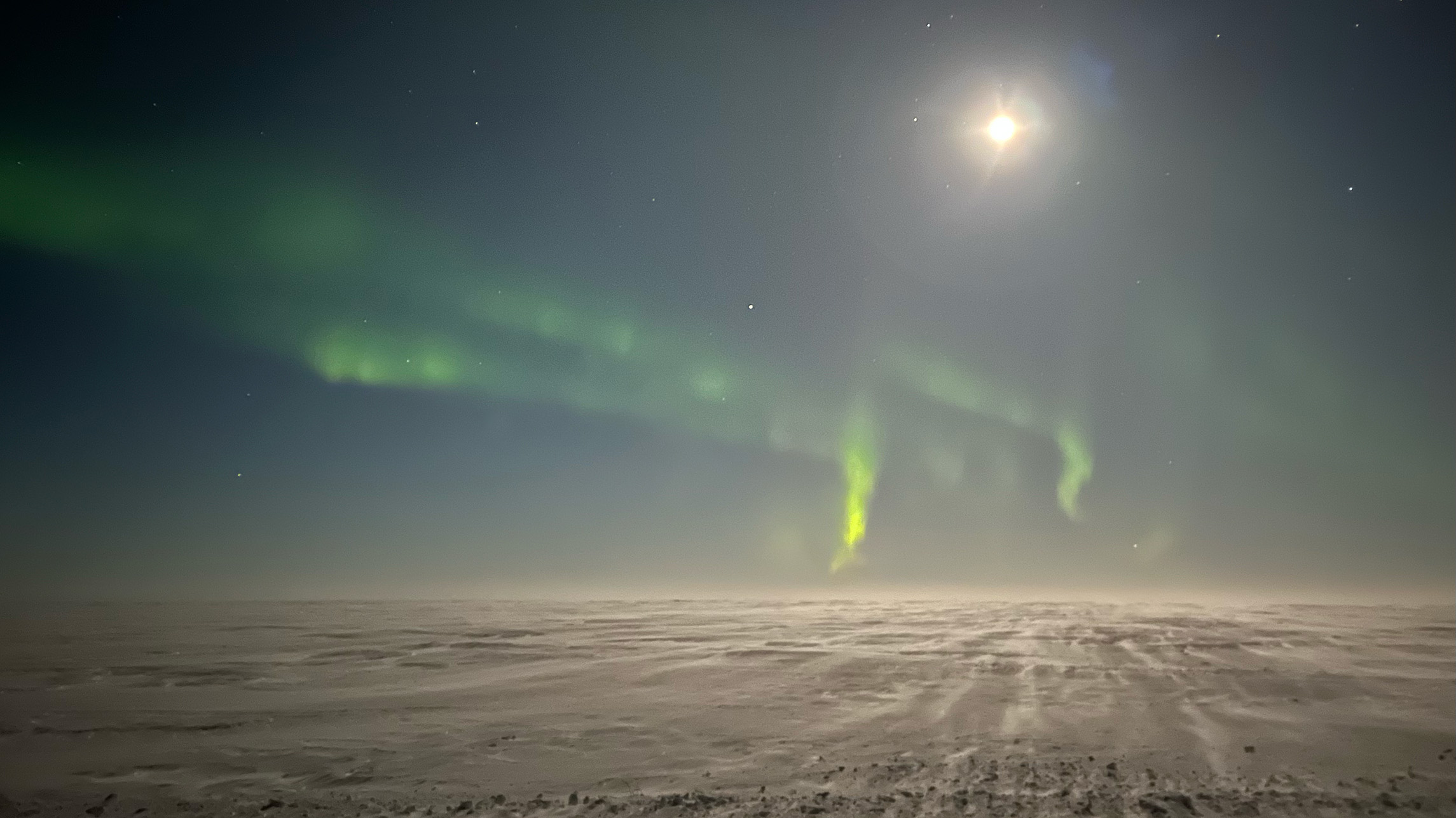 The image shows a landscape with a dark sky and a bright moon. There are wisps of the northern lights in green. The ground is covered in snow.