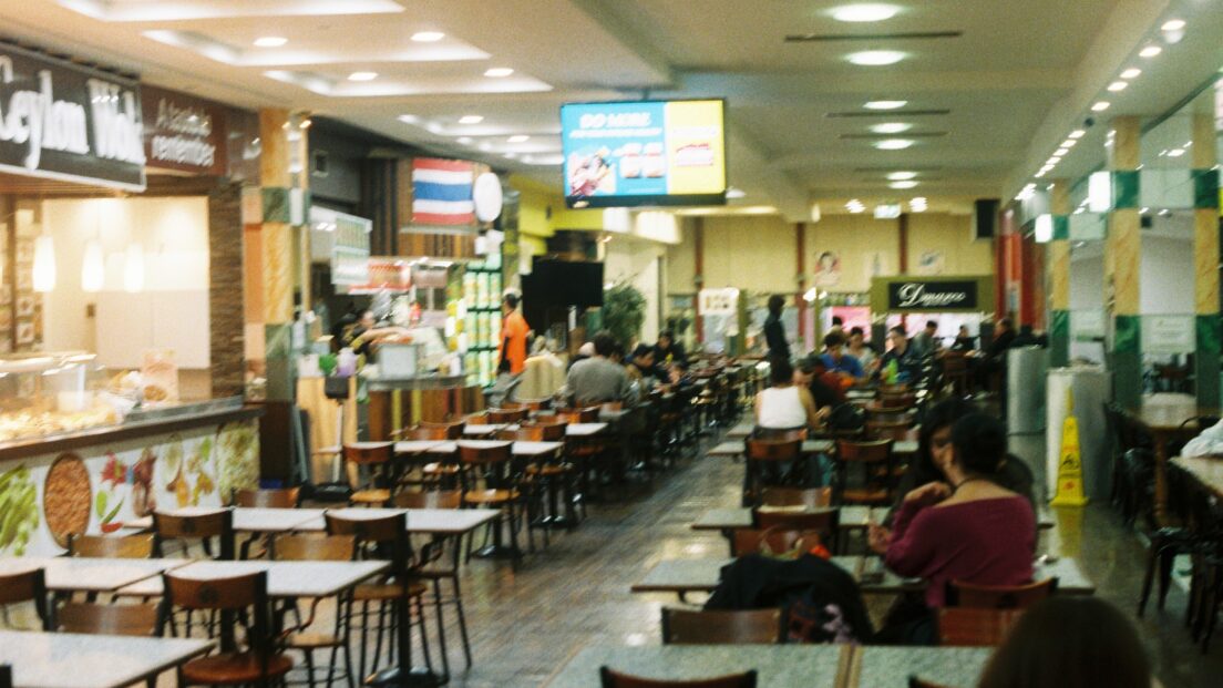 The image shows rows of tables and chairs inside the food court. There are food vendors to the left hand side.