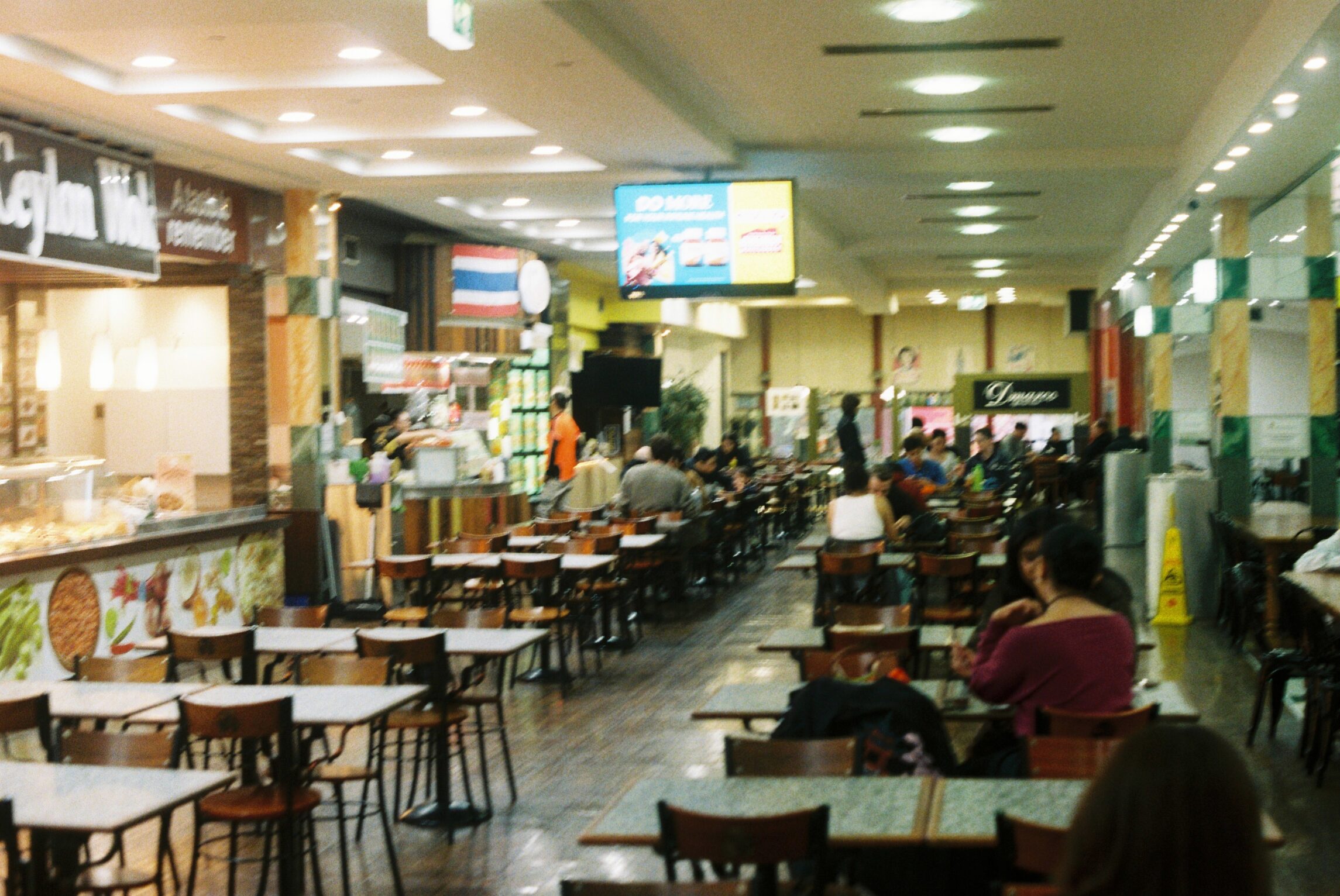 The image shows rows of tables and chairs inside the food court. There are food vendors to the left hand side.