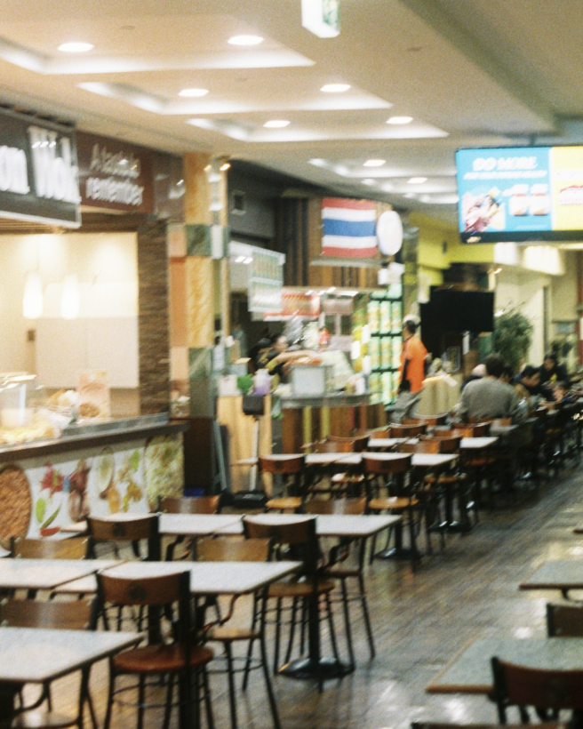 The image shows rows of tables and chairs inside the food court. There are food vendors to the left hand side.