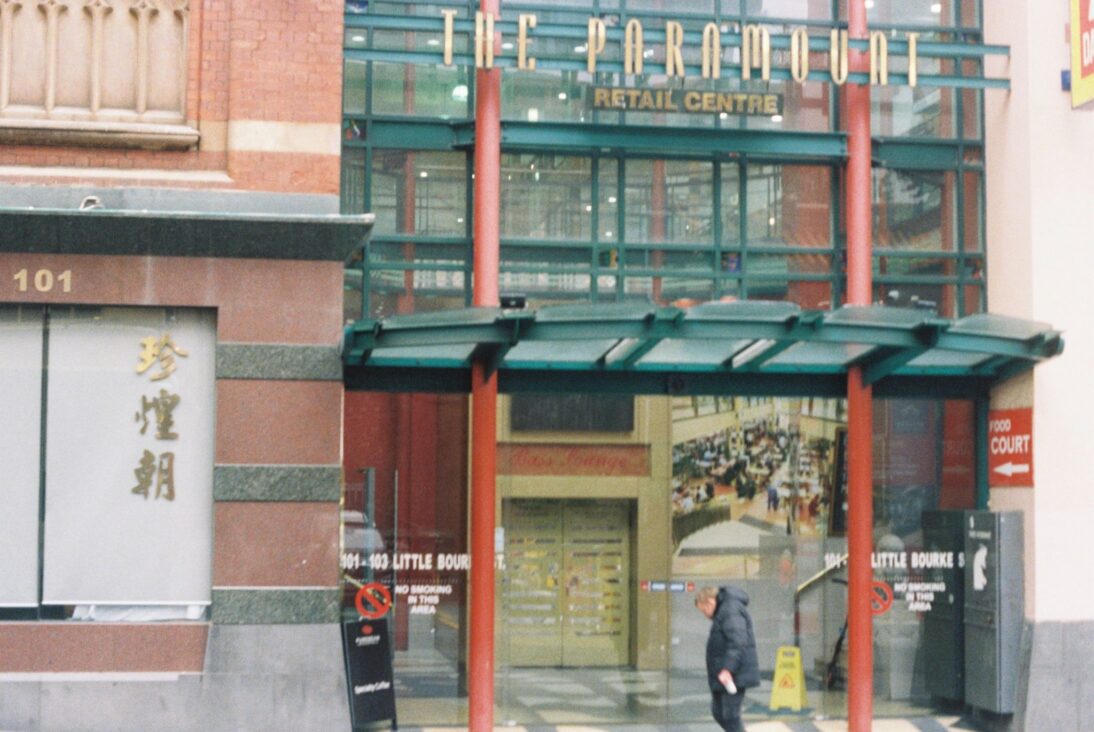 the image shows the entrance to the food court. There are red pillars with a green frame and there is a person stood outside with a grey jacket.