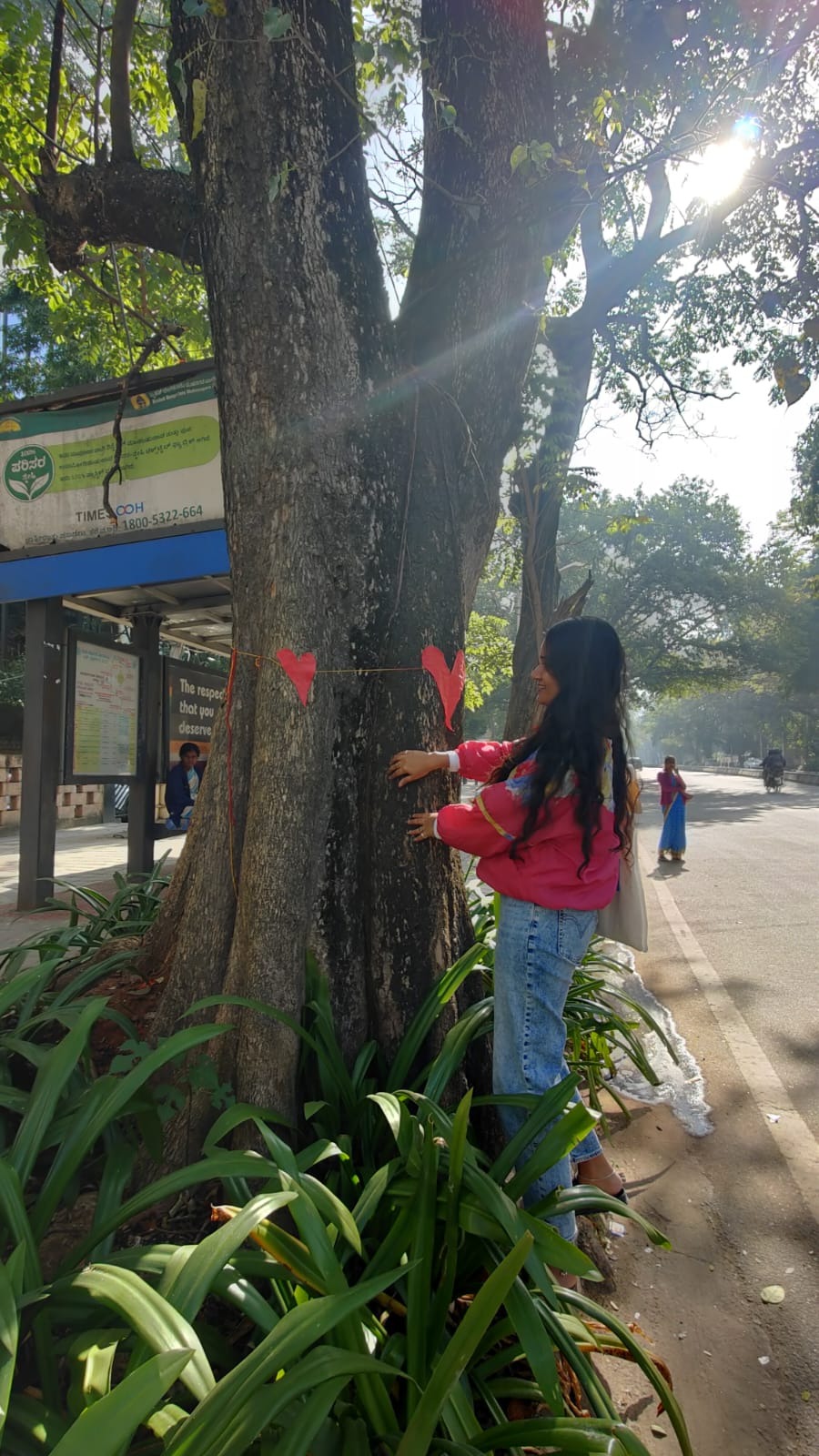 The image shows Divya standing next to a tree smiling. There are red hearts decorating the trunk of the tree.