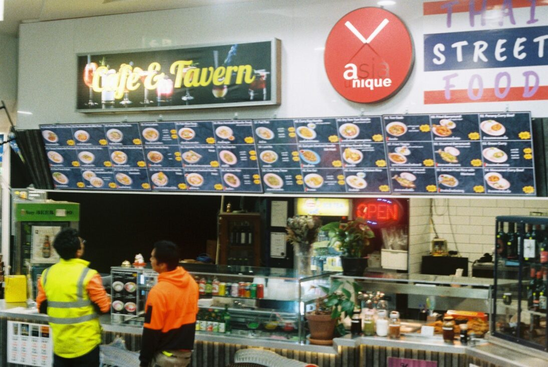 the image shows one of the food vendors inside the food court - there are two people stood in front, reading the signs. One wears a bright yellow high vis jacket and one wears an orange waterproof jacket.
