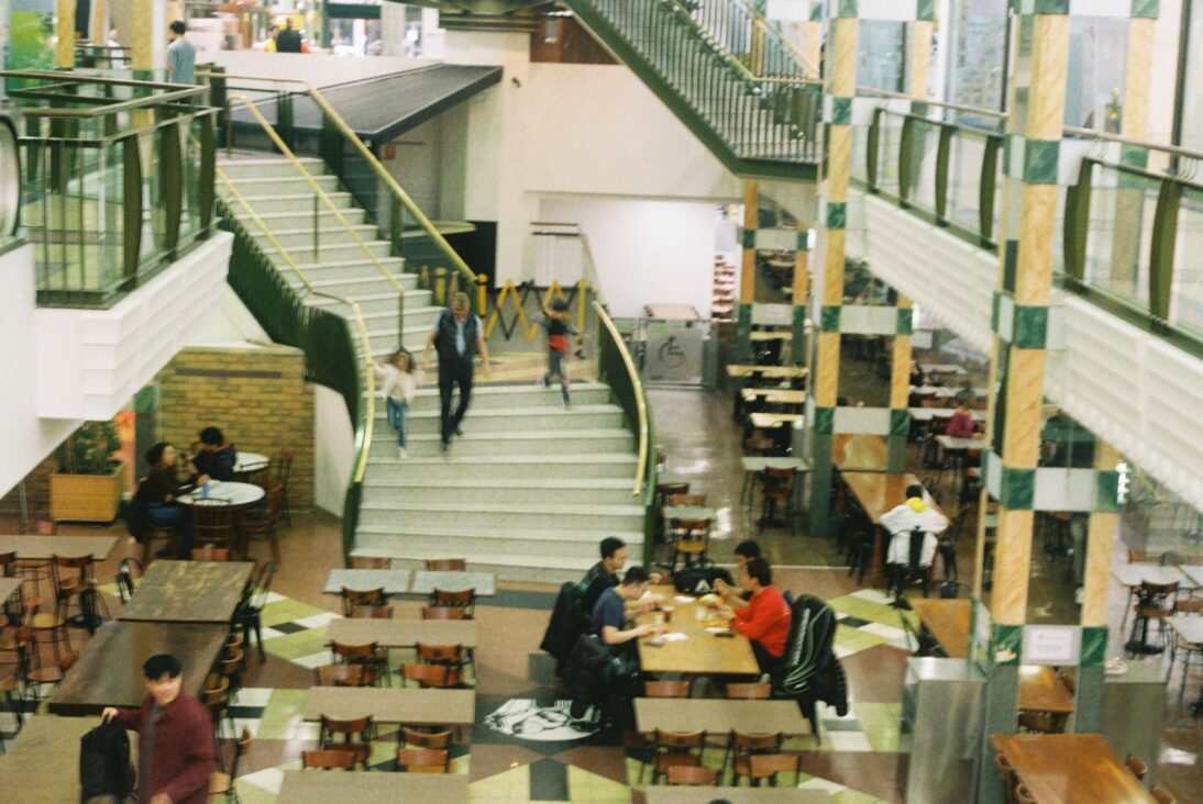 The image shows the inside of Paramount Food Court. There are rows of tables and a big staircase in the background. On the right hand side is a table of four people sat eating together