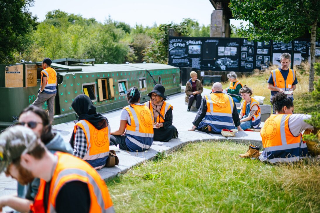 The image shows a group of people sat on the floor on a path next to the canal. There is a green houseboat to the left.