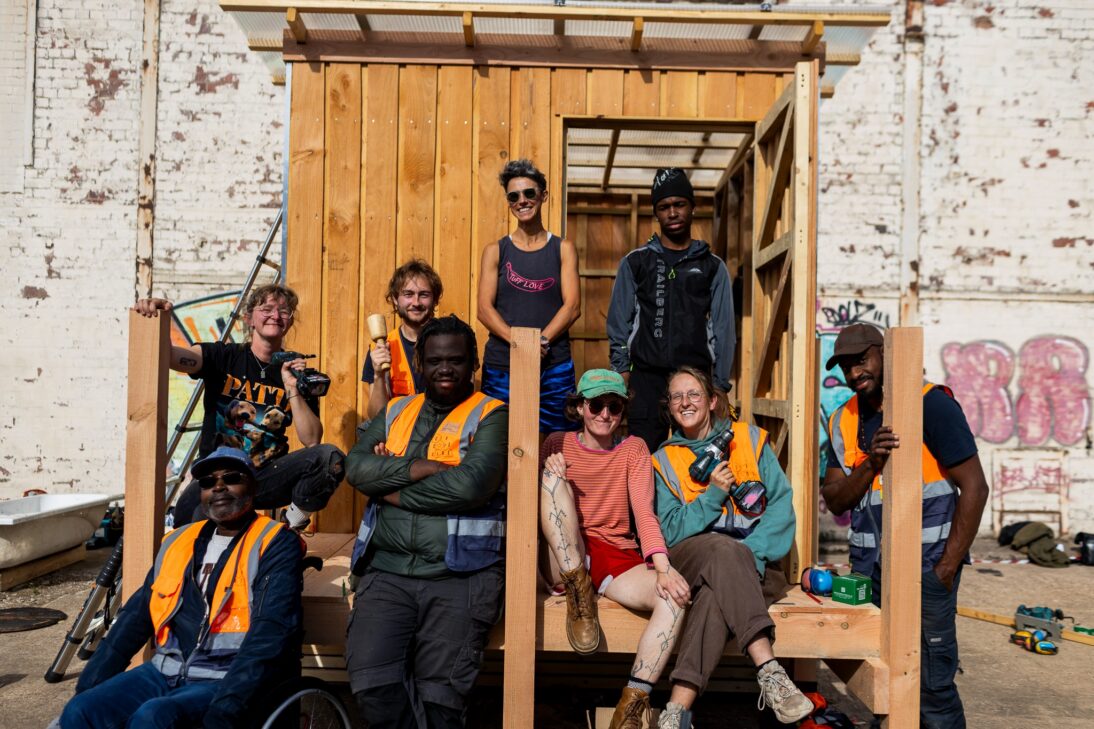 The image shows a group of people wearing orange high res jackets sat smiling at the camera. They are sat on a wooden structure that the are collectively building.