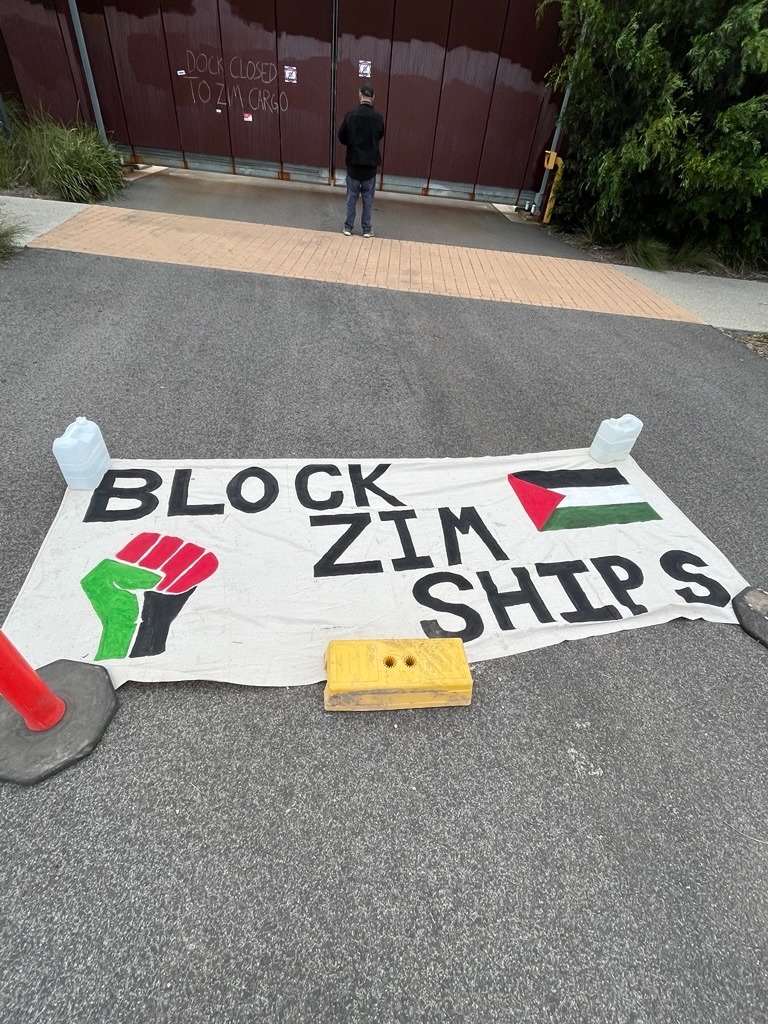 the image shows a white handpainted sign on the ground. There is a black power fist painted in the Palestinian colours on the left hand side and a palestinian flag on the right. The words read in black "Block SIM ships"