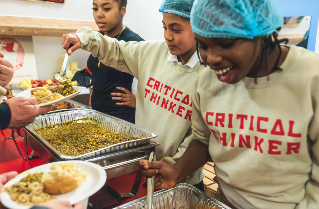the image shows young people serving food. they are wearing jumpers which say "critical thinker"