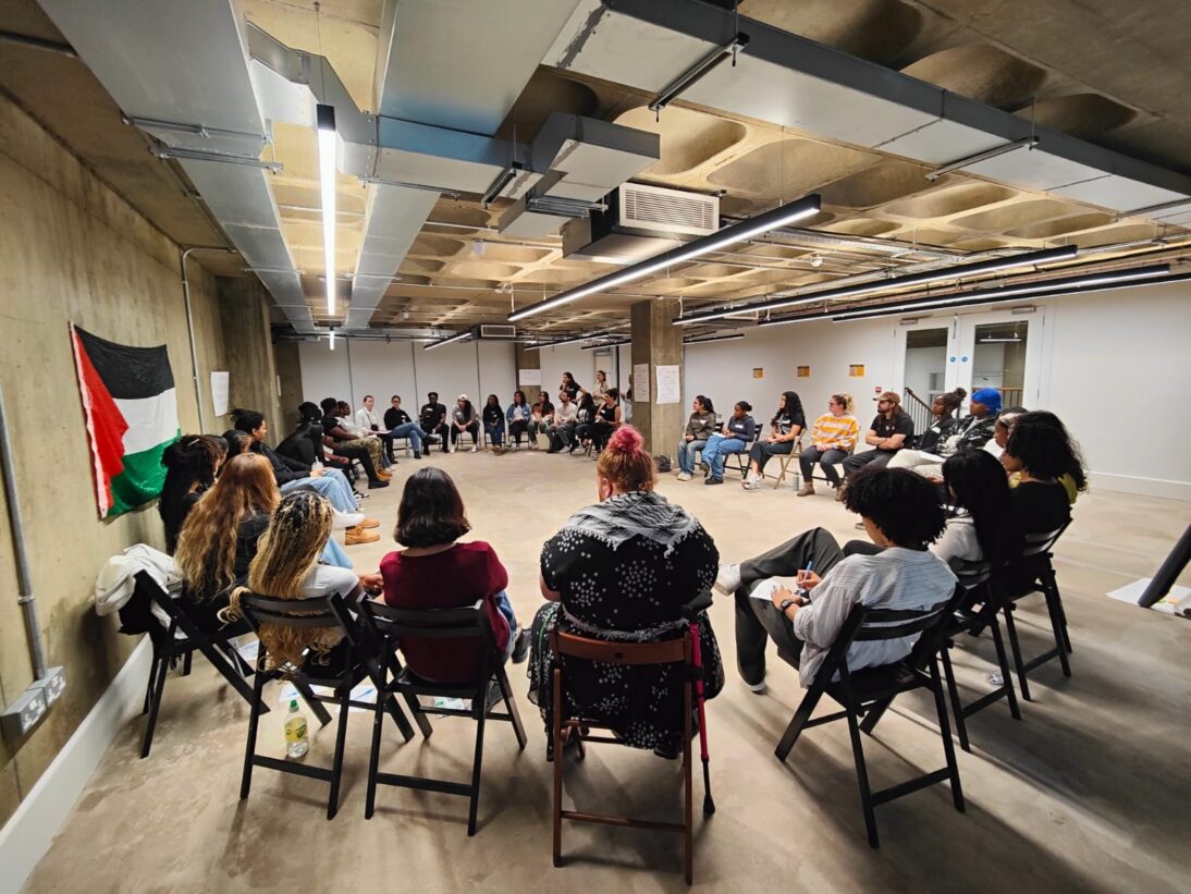 the image shows a group of young people sat on chairs in a circle shape. there is a palestinian flag on the wall