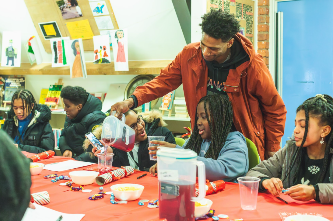 there is a group of young people sat at the table taking part in a workshop/group session. There is a member of staff pouring squash into cups