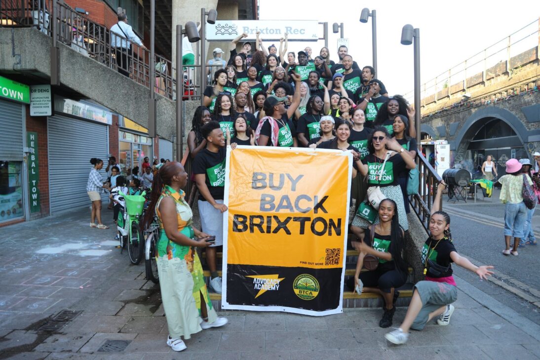 A group of young people stand on the stairs in support of the buy back brixton campaign. They are holding a big yellow sign and wear black t-shirts with a green sign saying buy back brixton