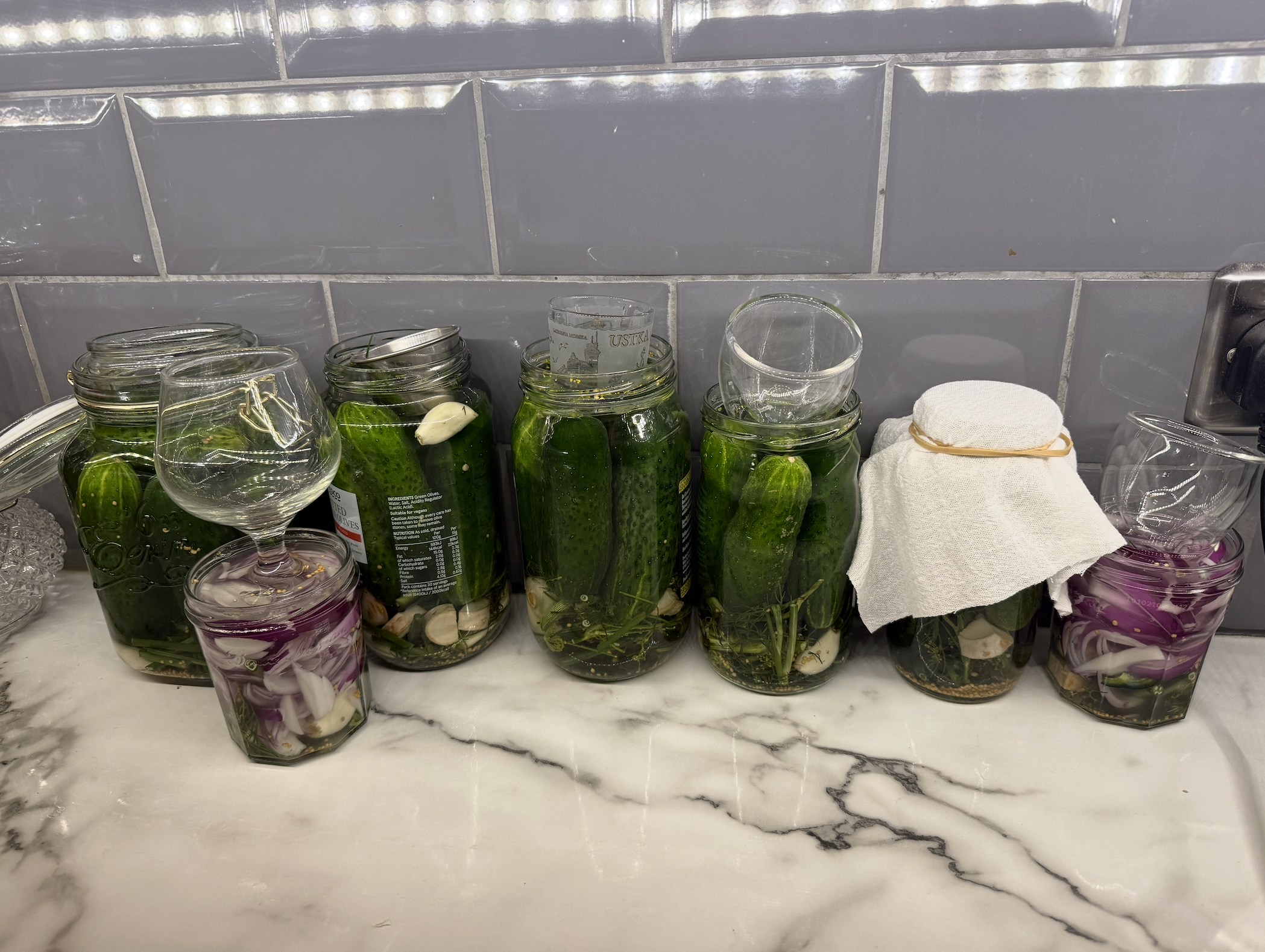 the image shows a row of jars of pickled gherkins on a marble working top