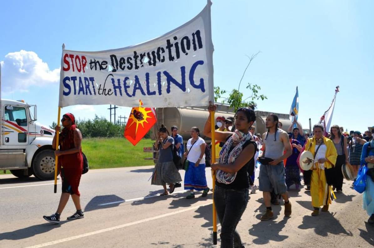 the image shows a group marching in protest. they are holding a banner that reads "stop the destruction, start the healing"
