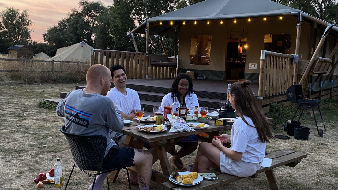 the image shows a group of friends sat around a picnic bench having dinner together