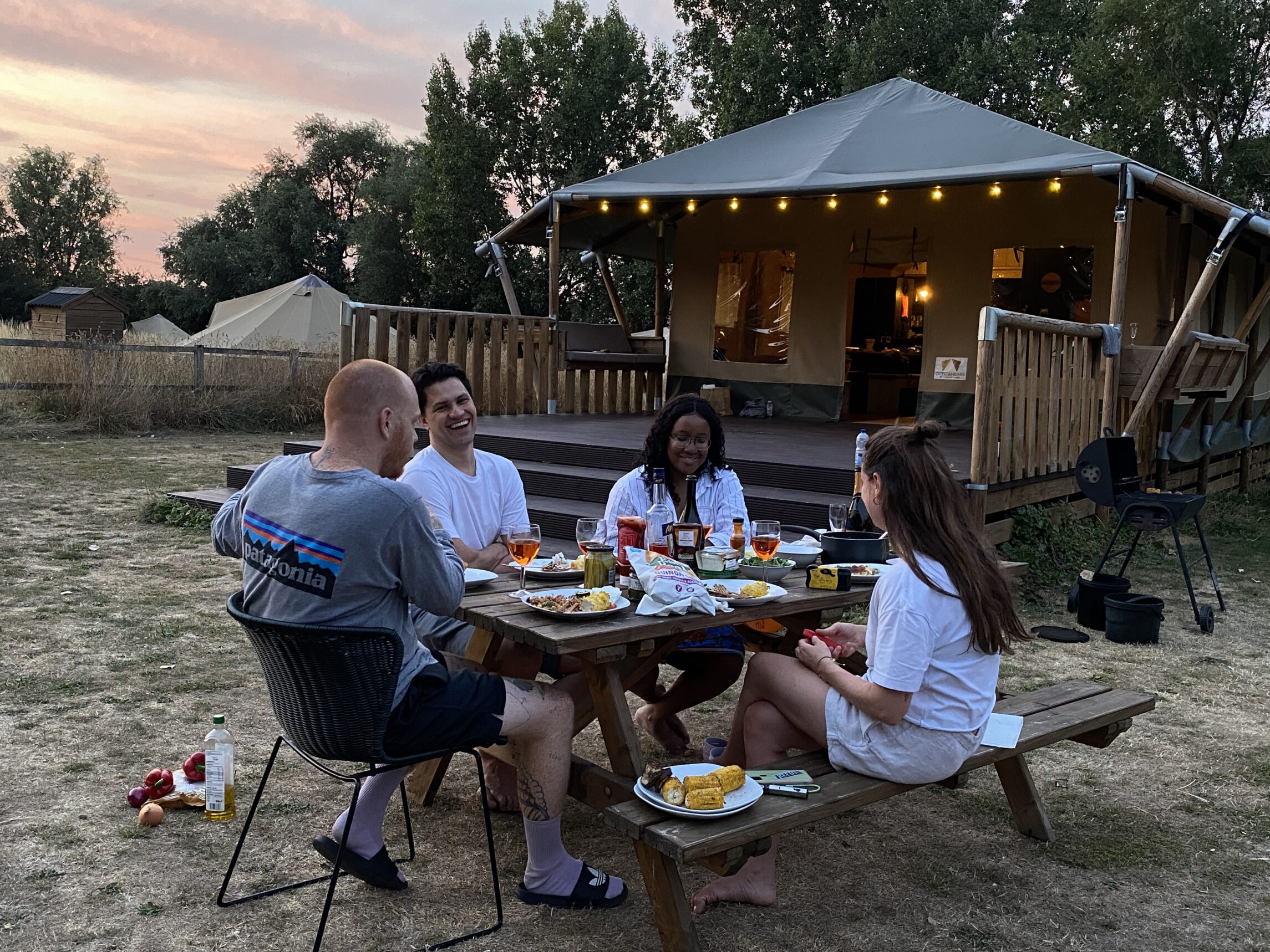 the image shows a group of friends sat around a picnic bench having dinner together