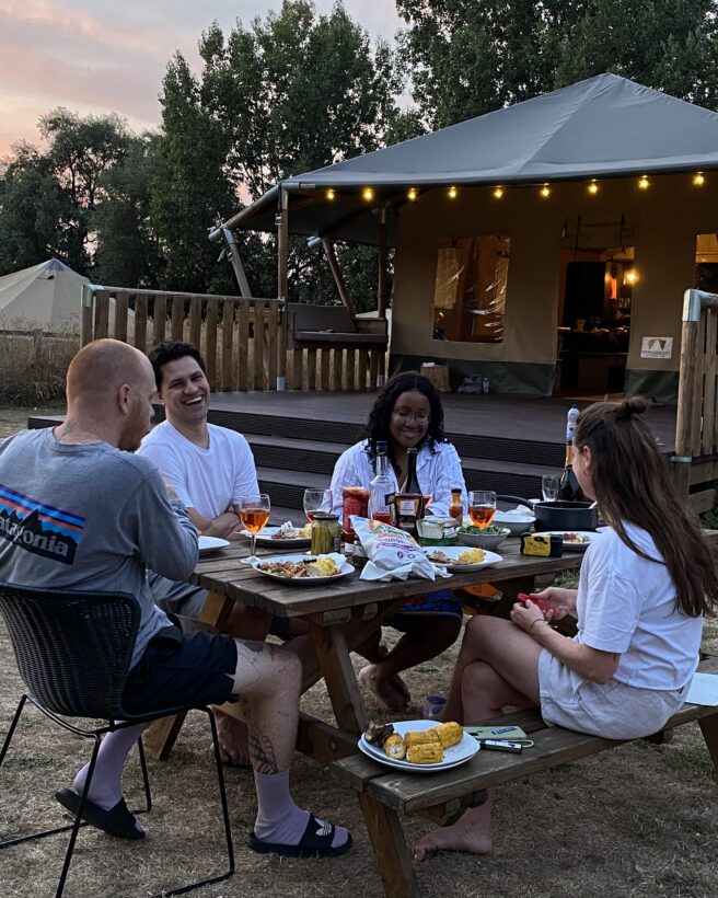 the image shows a group of friends sat around a picnic bench having dinner together